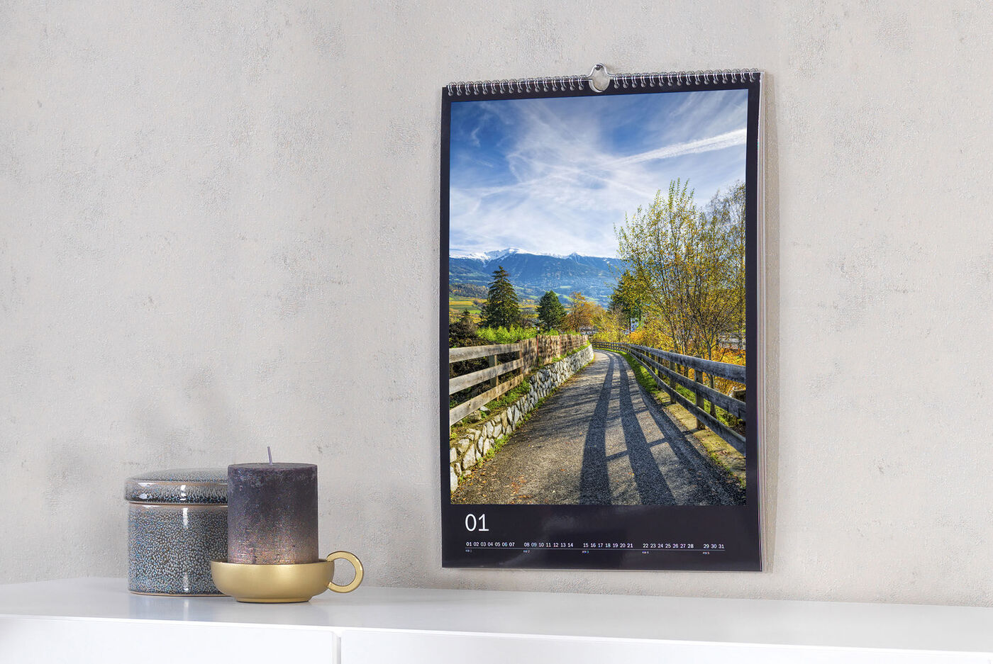 En vacker fotokalender hänger på en vägg. Bilden visar en målad stig mellan träd på en solig dag. En ljushållare, ett ljus och en kopp står på ett bord bredvid.
