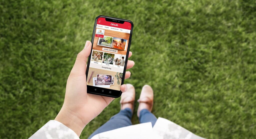En person håller en smartphone där CEWE-appen är öppen. Skärmen visar foton och designer. Bakgrunden är en grön gräsmatta.