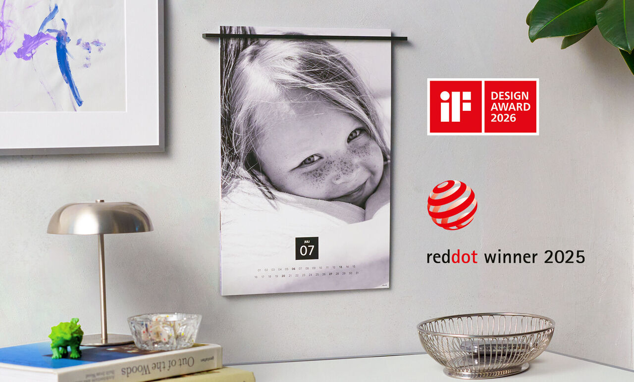 Väggkalender med ett leende barn och en vacker bakgrund. Kalendern visar juli månad och har red dot-designprisets logotyp.