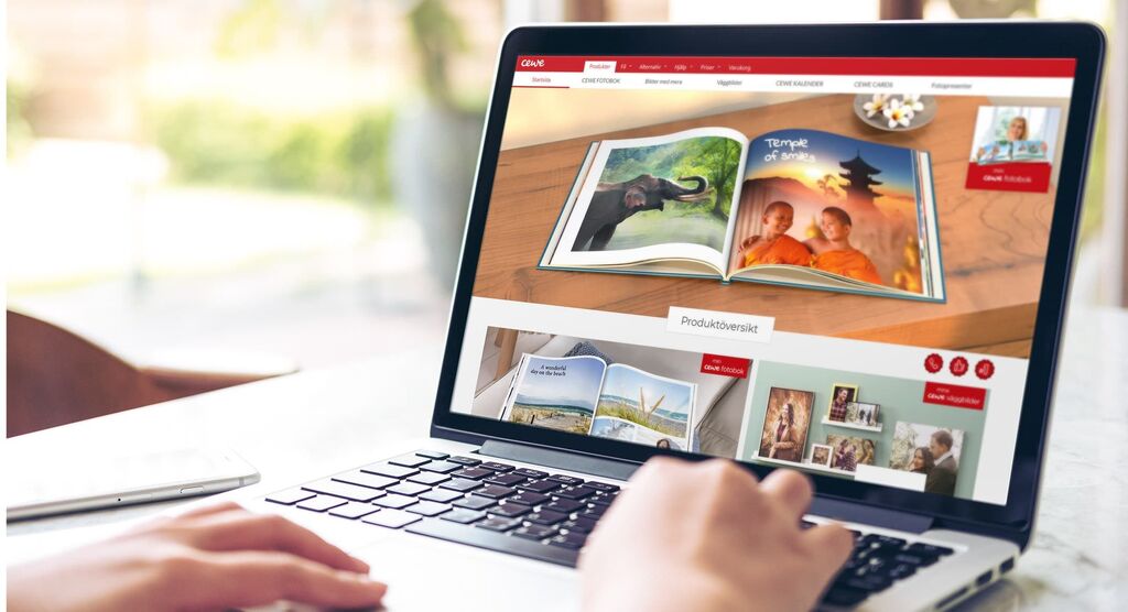 En person bläddrar entusiastiskt i ett öppet CEWE FOTOBOK på en laptop, omgivet av naturligt ljus.
