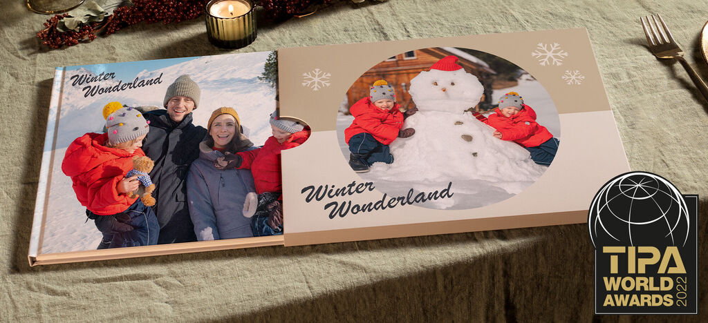 Två CEWE fotoböcker på ett bord, en visar en vinterfamiljescen med en snögubbe, den andra en lycklig familj i snön.