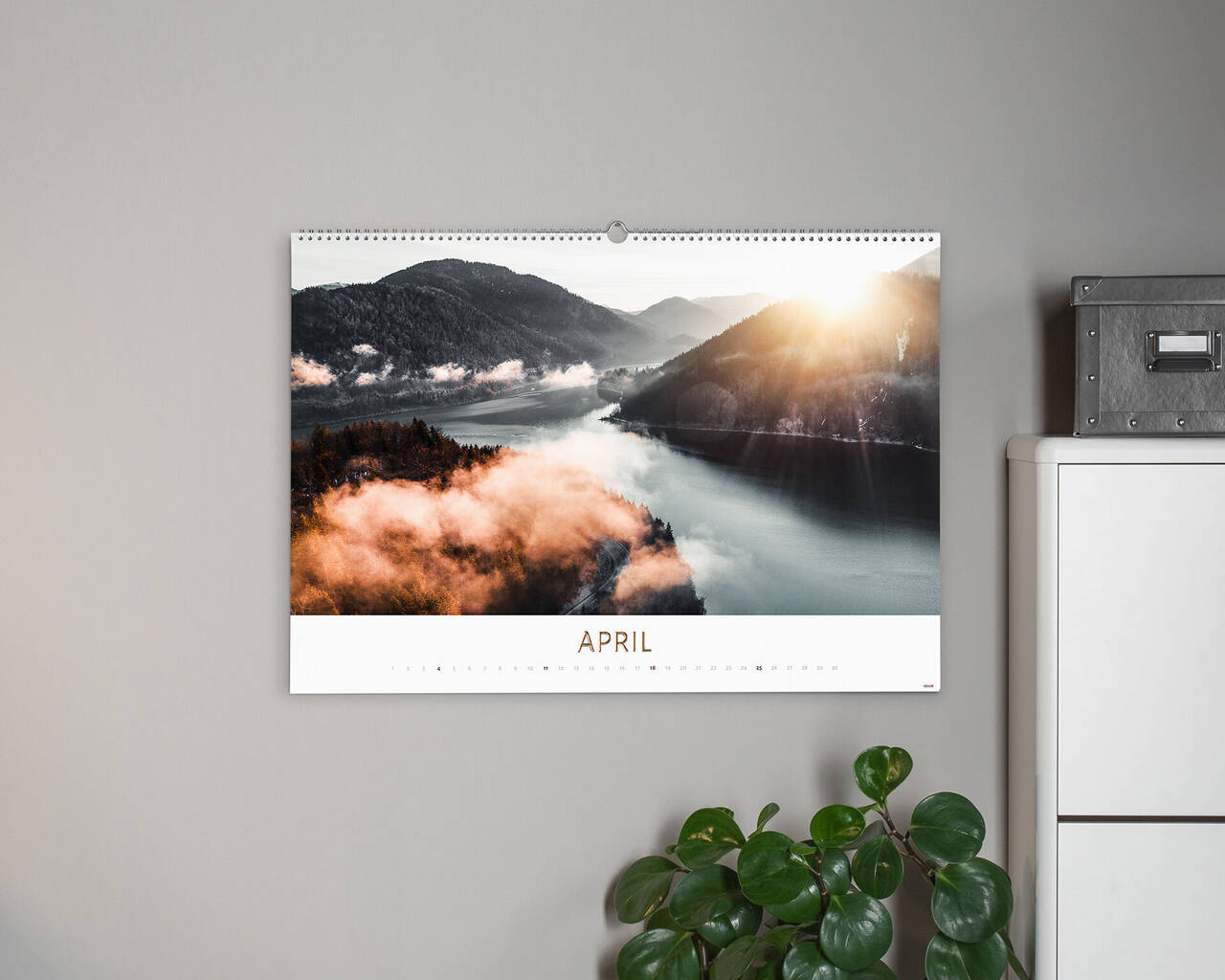 Vacker fotokalender med landskapsbild för april. Kalendern visar en soluppgång över en dimmig sjö omgiven av gröna berg.