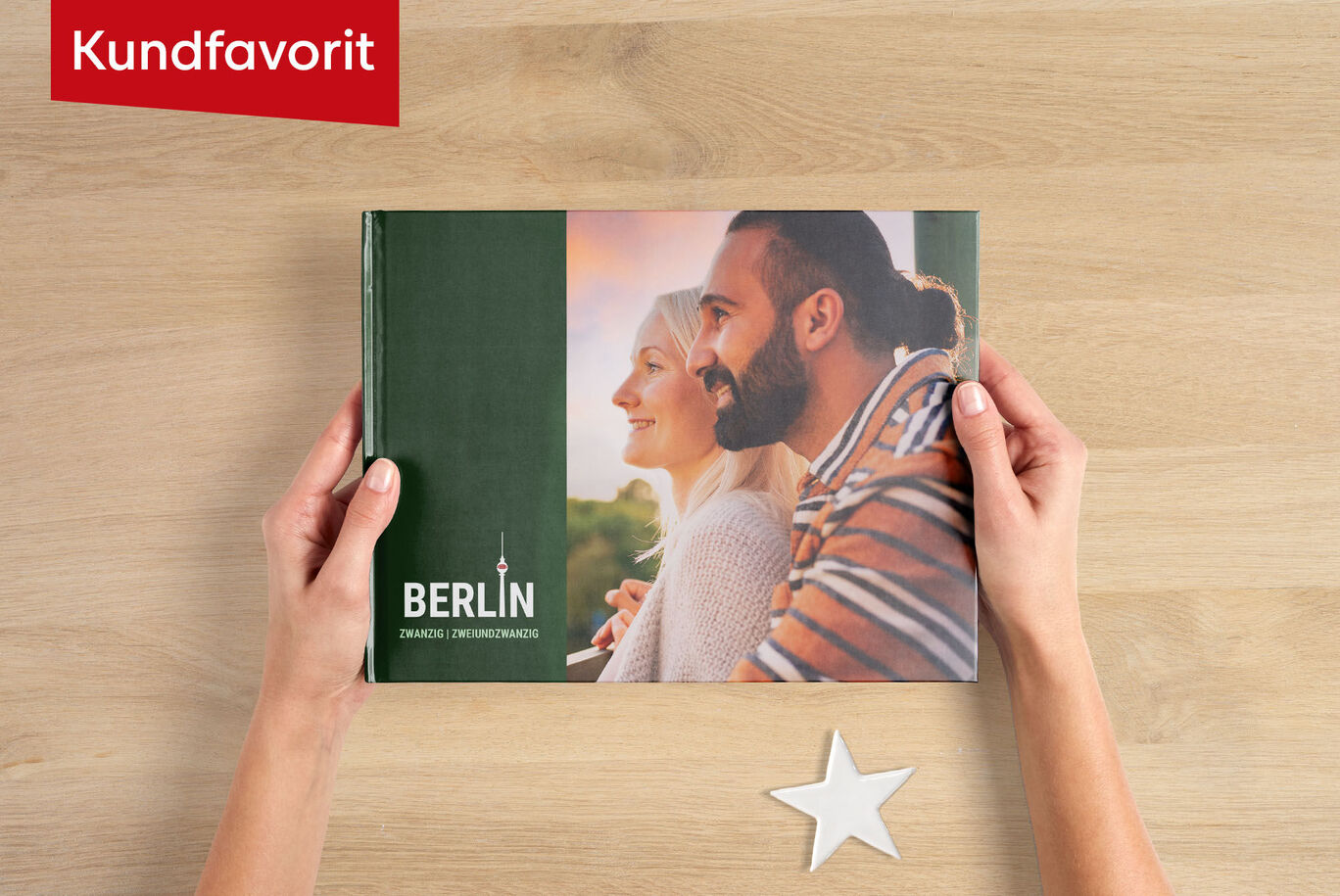 En person håller en CEWE FOTOBOOK med titeln "Berlin" på en träyta omgiven av stjärndekoration.