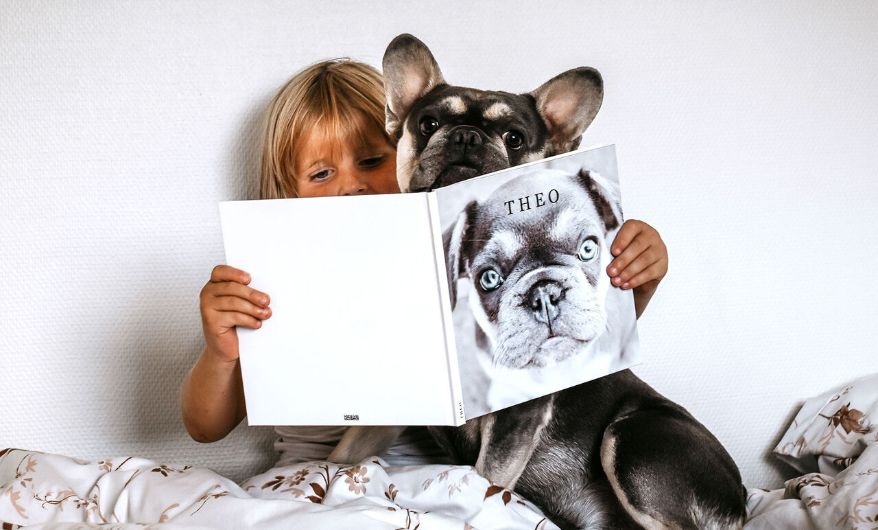 Ett barn håller en bok med titeln 'THEO' medan en hund sitter bredvid och ser på betraktaren.