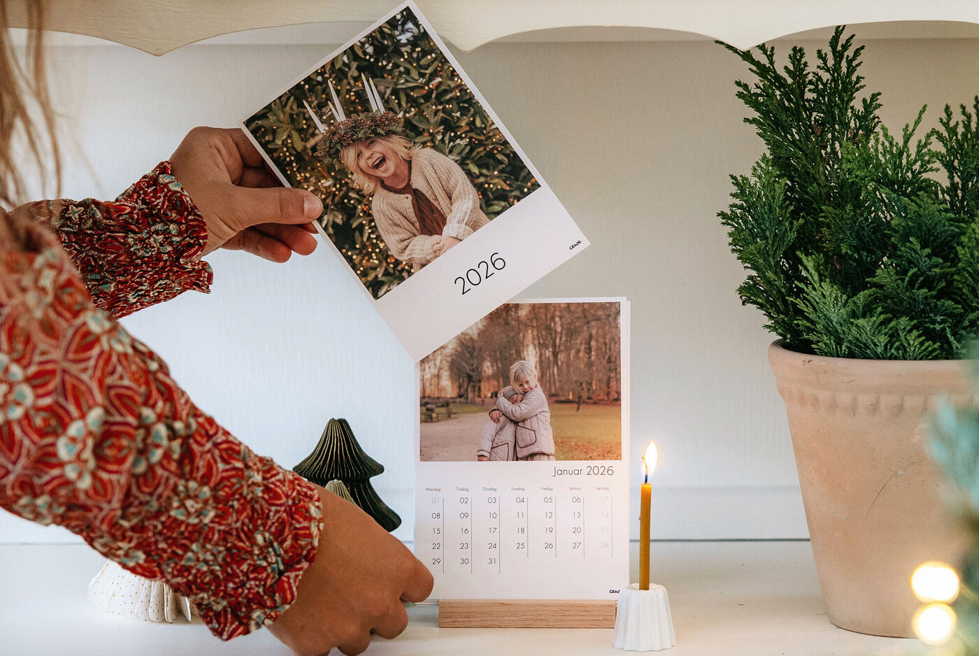 En person håller en uppställd fotokalender med bilder och år 2025 på ett bord. Ett ljus brinner bredvid en julgran.