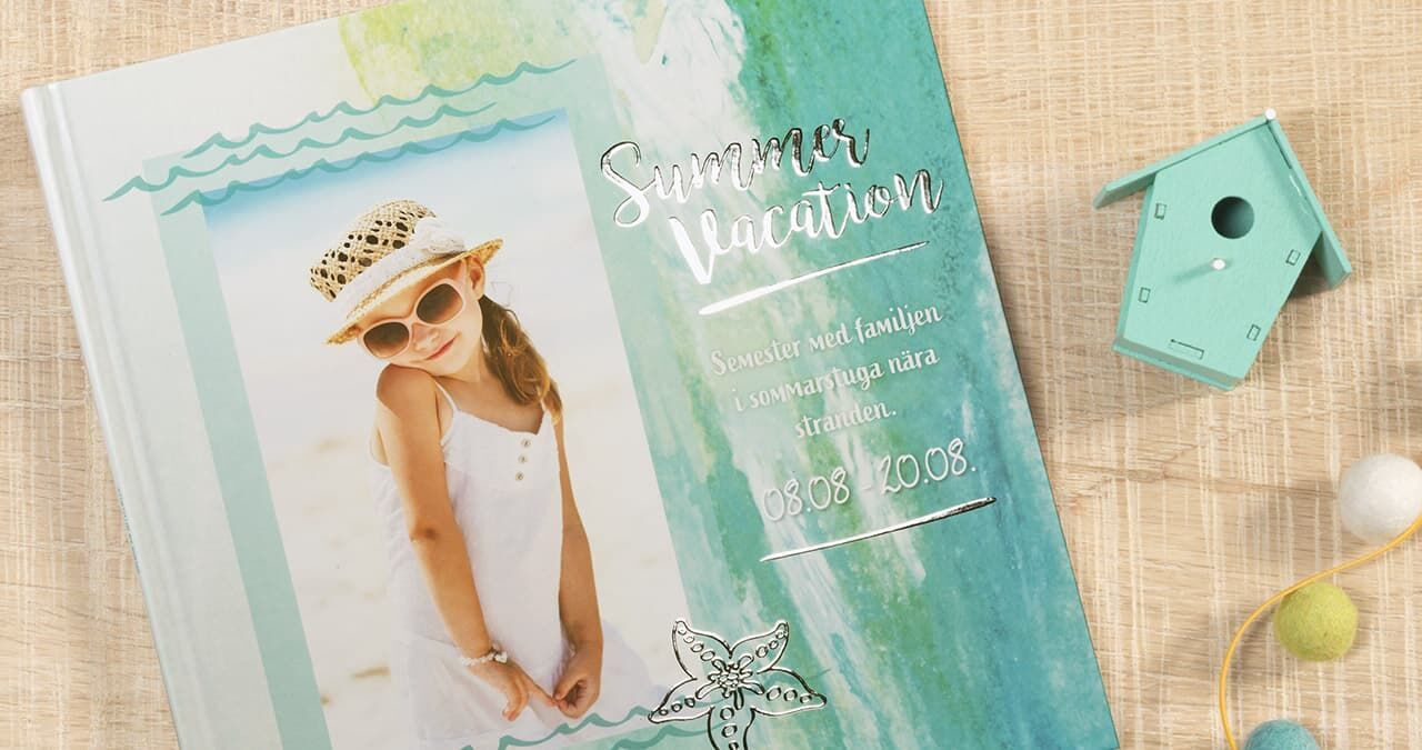 Ett vackert designat CEWE Fotobok med titeln 'Summer Vacation' och en maritim bakgrund. Det ligger på ett ljust bord, omgiven av fågelhusdekoration och färgglada pompons.