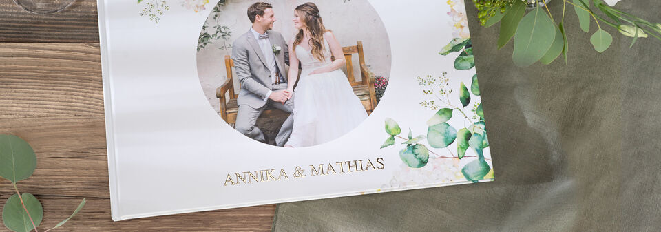 En vacker CEWE fotobok med titeln Annika & Matthias på ett träbord, omgiven av gröna blad.